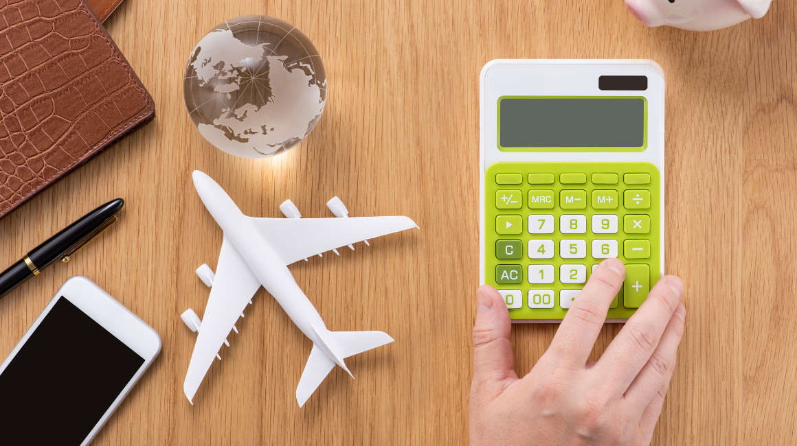 Planowanie budżetu podróży — ręka na kalkulatorze, obok miniaturowy samolot, kula ziemska, telefon, portfel i długopis na drewnianym biurku